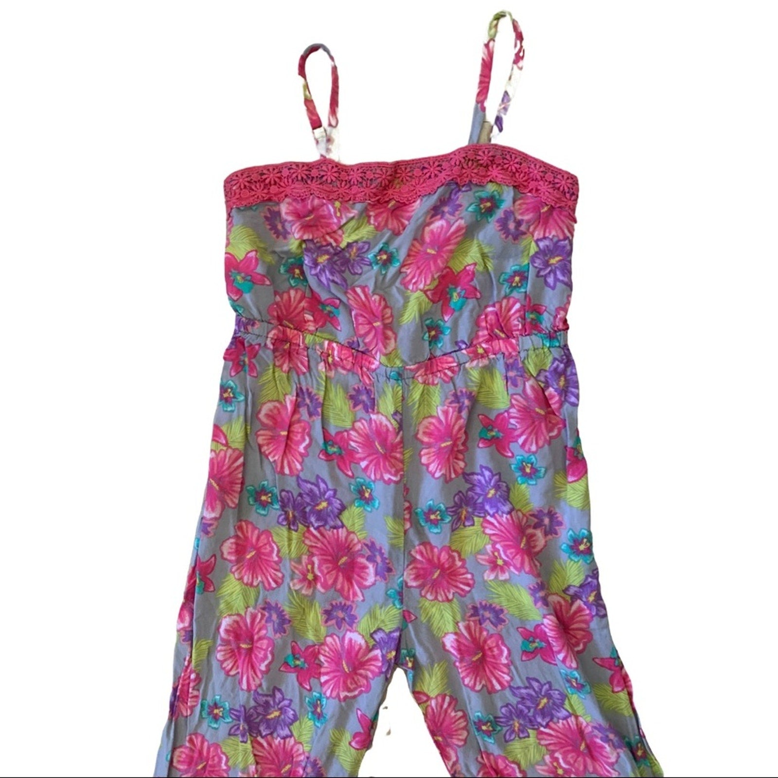 A sleeveless floral jumpsuit with thin shoulder straps, featuring a vibrant pink, purple, and green floral pattern on a light blue-gray base, with a pink lace-trimmed neckline.
