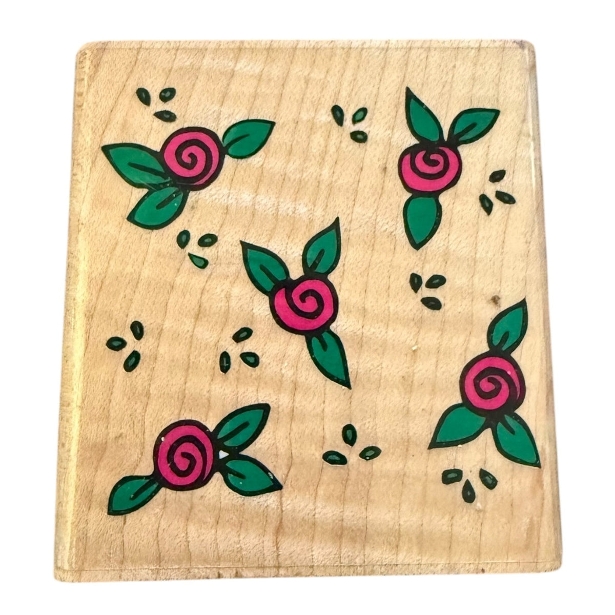 A square wooden block with pattern of pink roses with green leaves and small black dots on a light wood grain background.