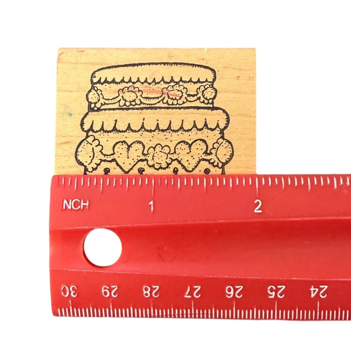 A wooden rubber stamp featuring a black ink illustration of a two-tiered cake with floral decorations, shown next to a red plastic ruler for scale on a white background.