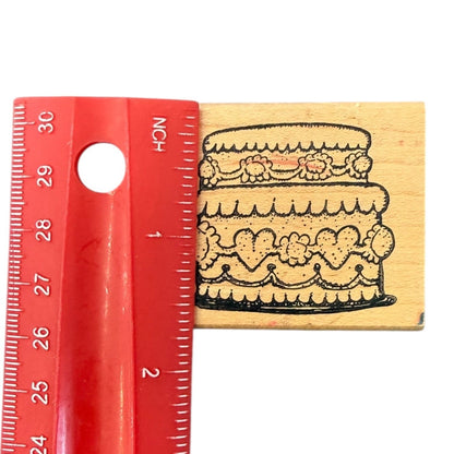 A wooden rubber stamp featuring a black ink design of a two-tiered cake with floral decorations, shown next to a red plastic ruler for scale on a white background.