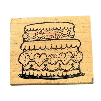 A square wooden rubber stamp featuring a black ink illustration of a three-tiered decorated cake, shown on a plain white background.