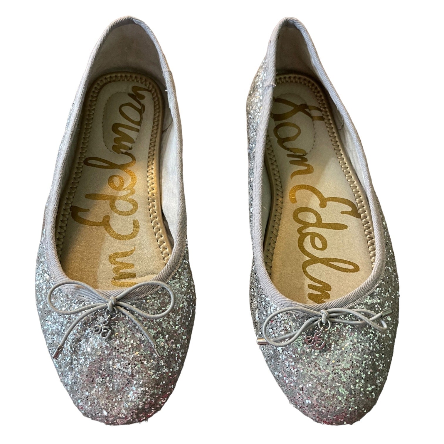 A pair of silver glitter ballet flats with a small bow on the toe, shown from both the top and side views against a white background. The insole features the 'Sam Edelman' brand name in gold script.