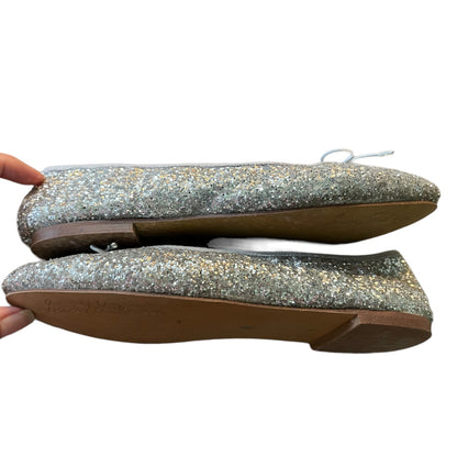 A pair of glittery silver flat shoes with brown soles, held by a hand against a plain white background.