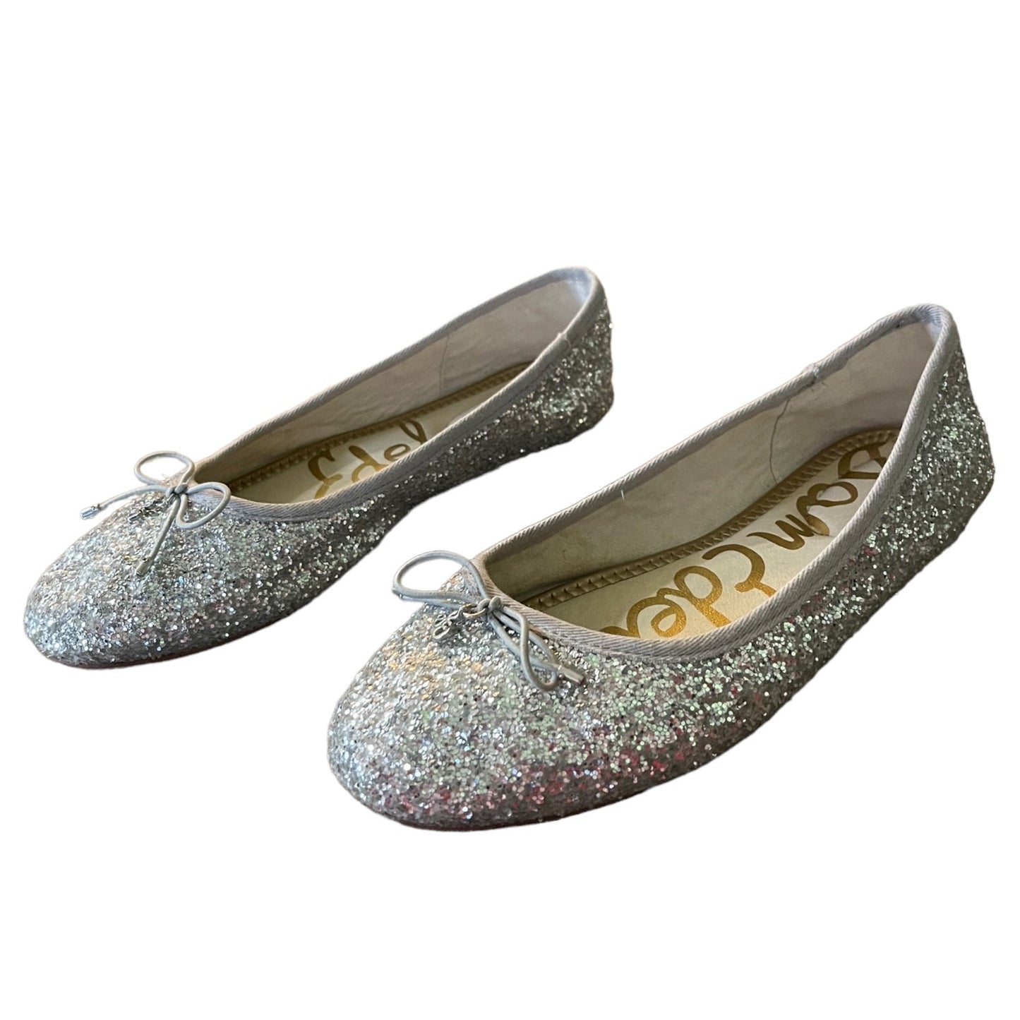 A pair of girl's flat shoes covered in silver glitter, featuring a small silver bow on the toe and a visible 'Sam Edco' brand name printed on the insole.