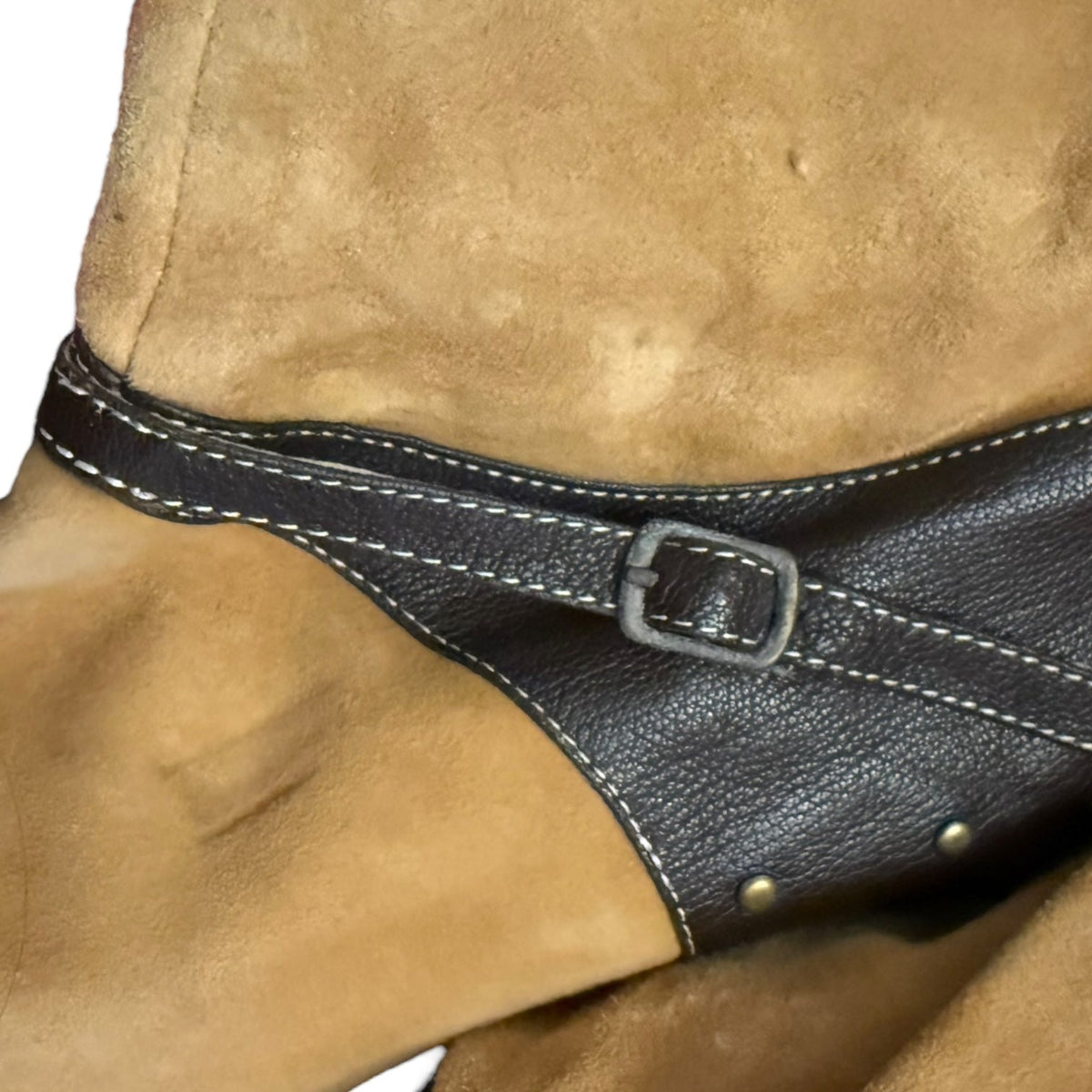 Close-up of a tan suede boot with faux leather strap and buckle closure, showing white contrast stitching and metal studs.