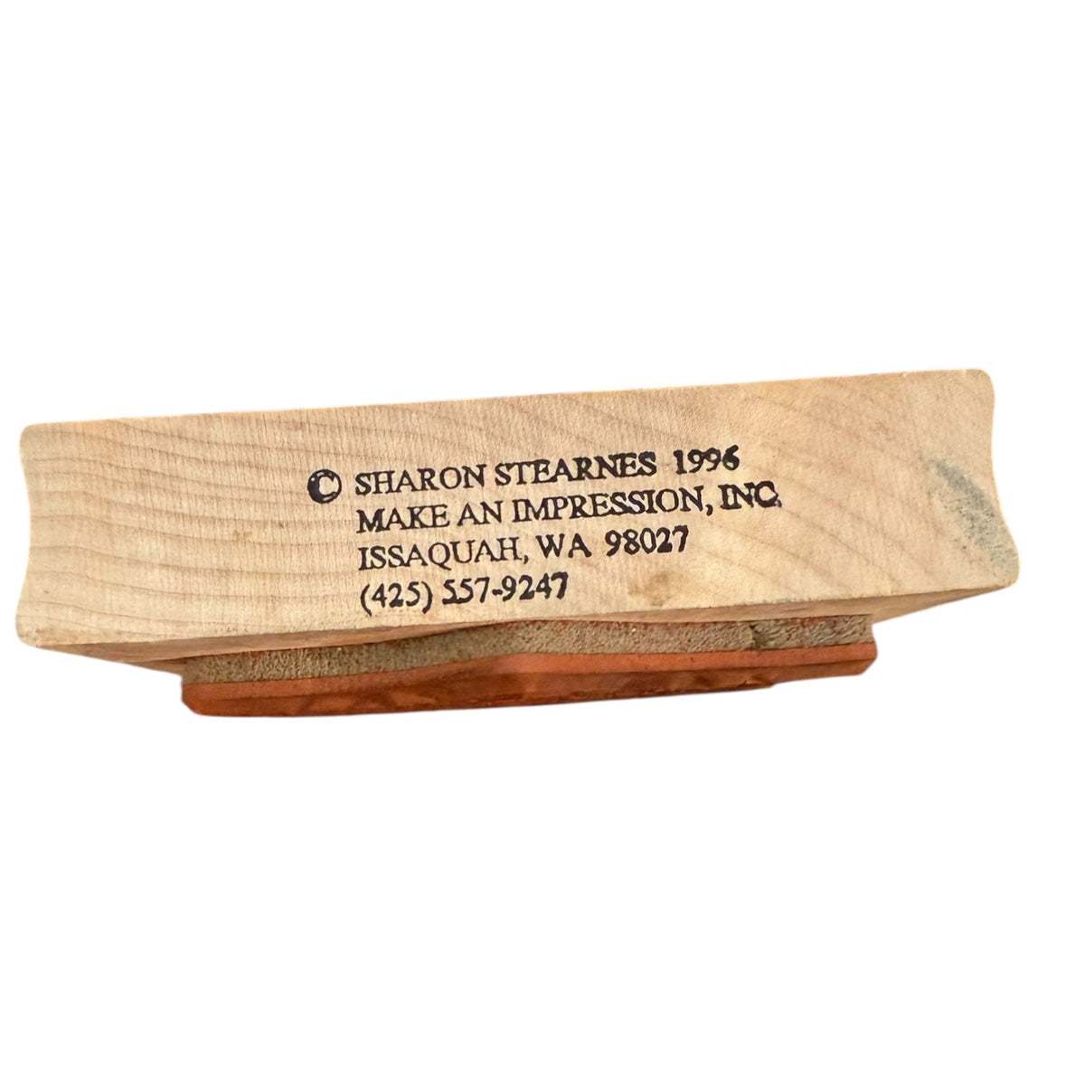 A custom wooden stamp block with a  wood, featuring engraved text that reads '© SHARON STEARNES 1996 MAKE AN IMPRESSION, INC. ISSAQUAH, WA 98027 (425) 557-9247' on a plain white background.