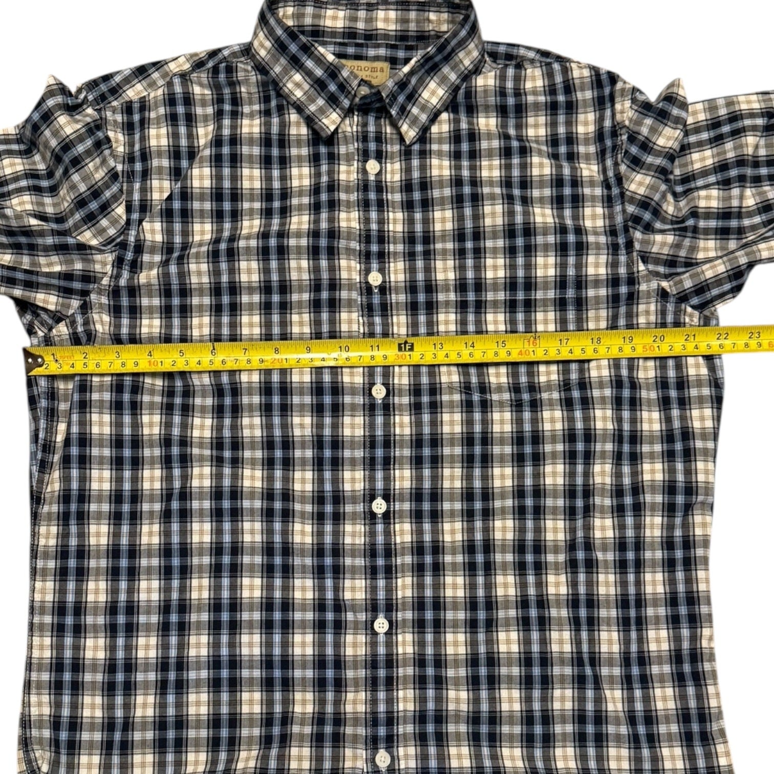 A short-sleeve plaid button-up shirt laid flat on a white background, with a yellow tape measure stretched horizontally across the chest to show the width.