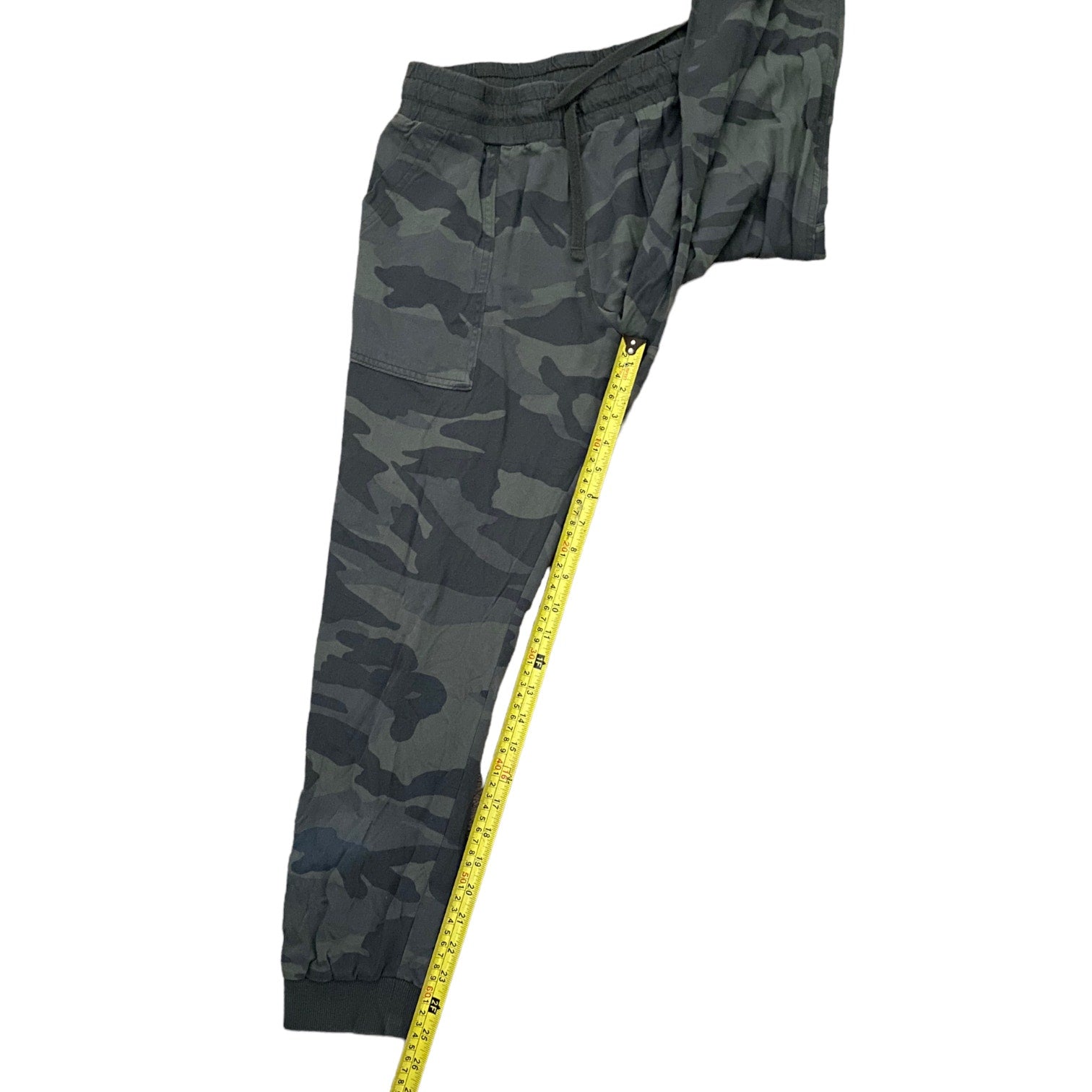 Dark camouflage jogger pants shown on a white background with a yellow measuring tape running down the side to indicate length.