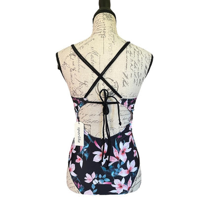 A women's one-piece swimsuit with a black base and a pink, blue, and white tropical floral print, shown from the back on a vintage-style paper dress form mannequin. The back features criss-cross black straps and a tie closure.