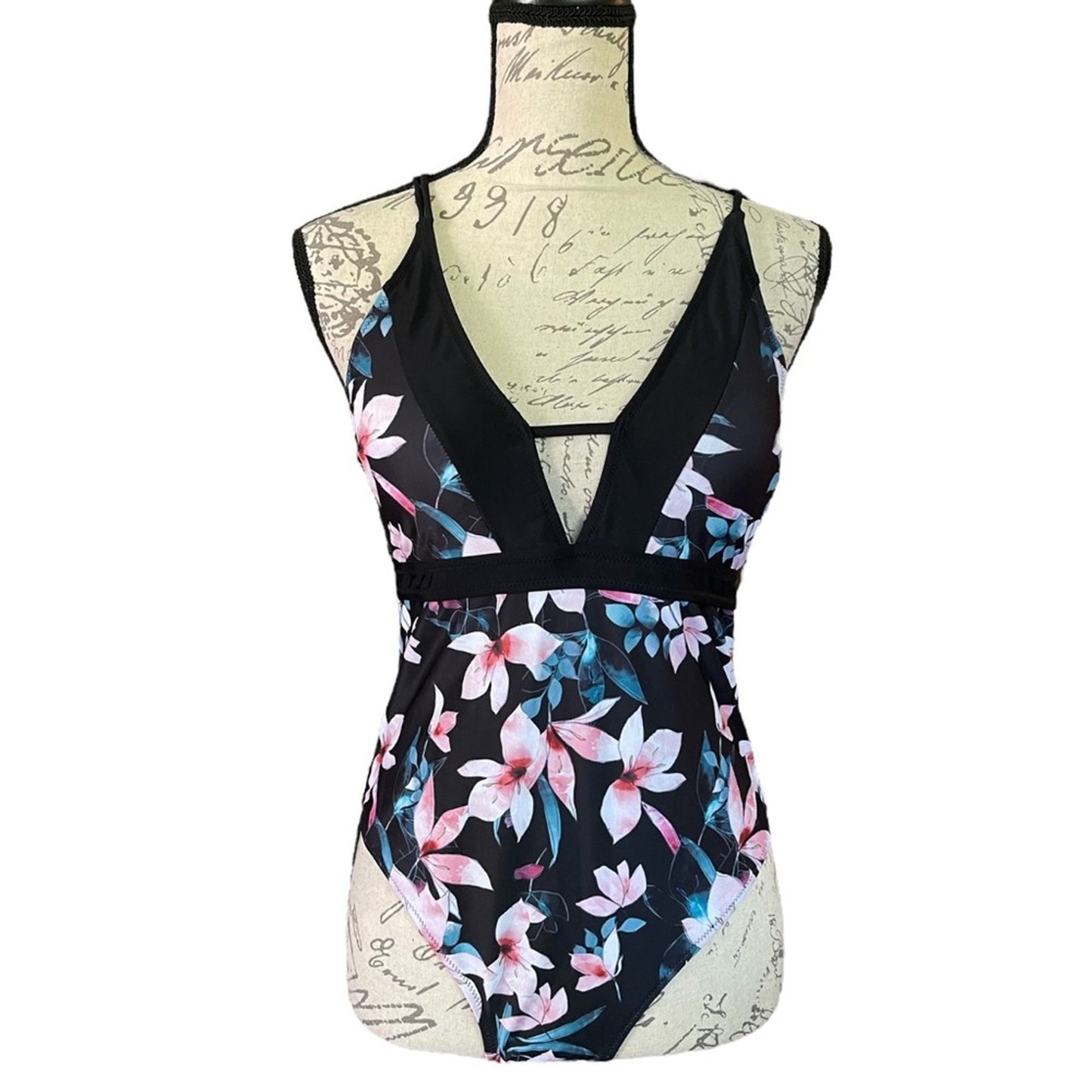A one-piece swimsuit displayed on a mannequin torso against a plain white background. The swimsuit has a black base with a pink, blue, and white tropical floral print, featuring a deep V-neckline with a keyhole cutout and a thin black waistband.