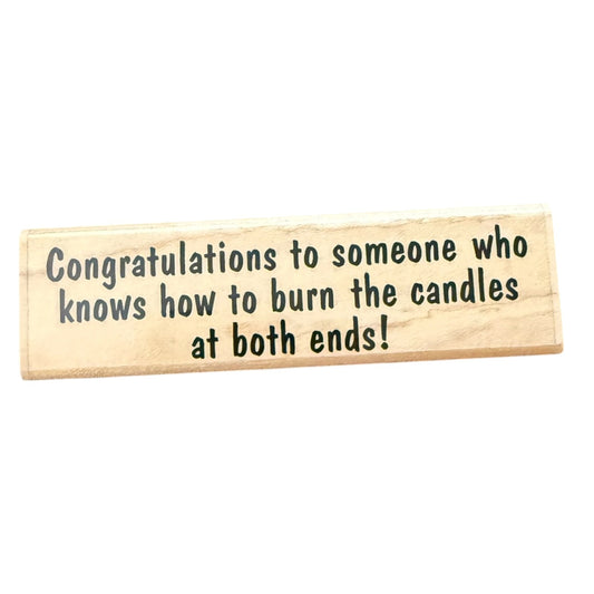 A rectangular light wood rubber stamp with black printed text that reads, 'Congratulations to someone who knows how to burn the candles at both ends!' on a plain white background.