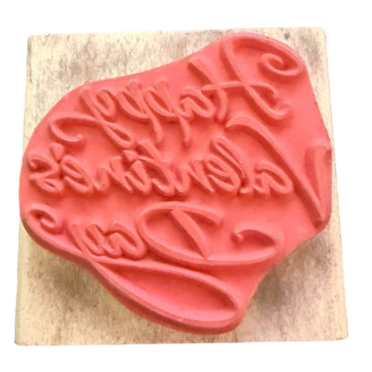 A pink rubber stamp with the words 'Happy Valentine's Day' in cursive, mounted on a square light wood block against a white background.