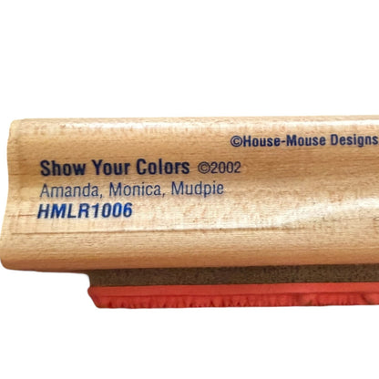 A close-up of a wooden stamp with blue printed text reading 'Show Your Colors ©2002 Amanda, Monica, Mudpie HMLR1006' and '©House-Mouse Designs', with a red rubber stamp face partially visible below on a white background.