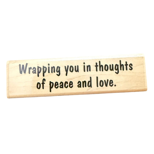 A rectangular light wood rubber stamp with black printed text that reads, 'Wrapping you in thoughts of peace and love.' on a plain white background.