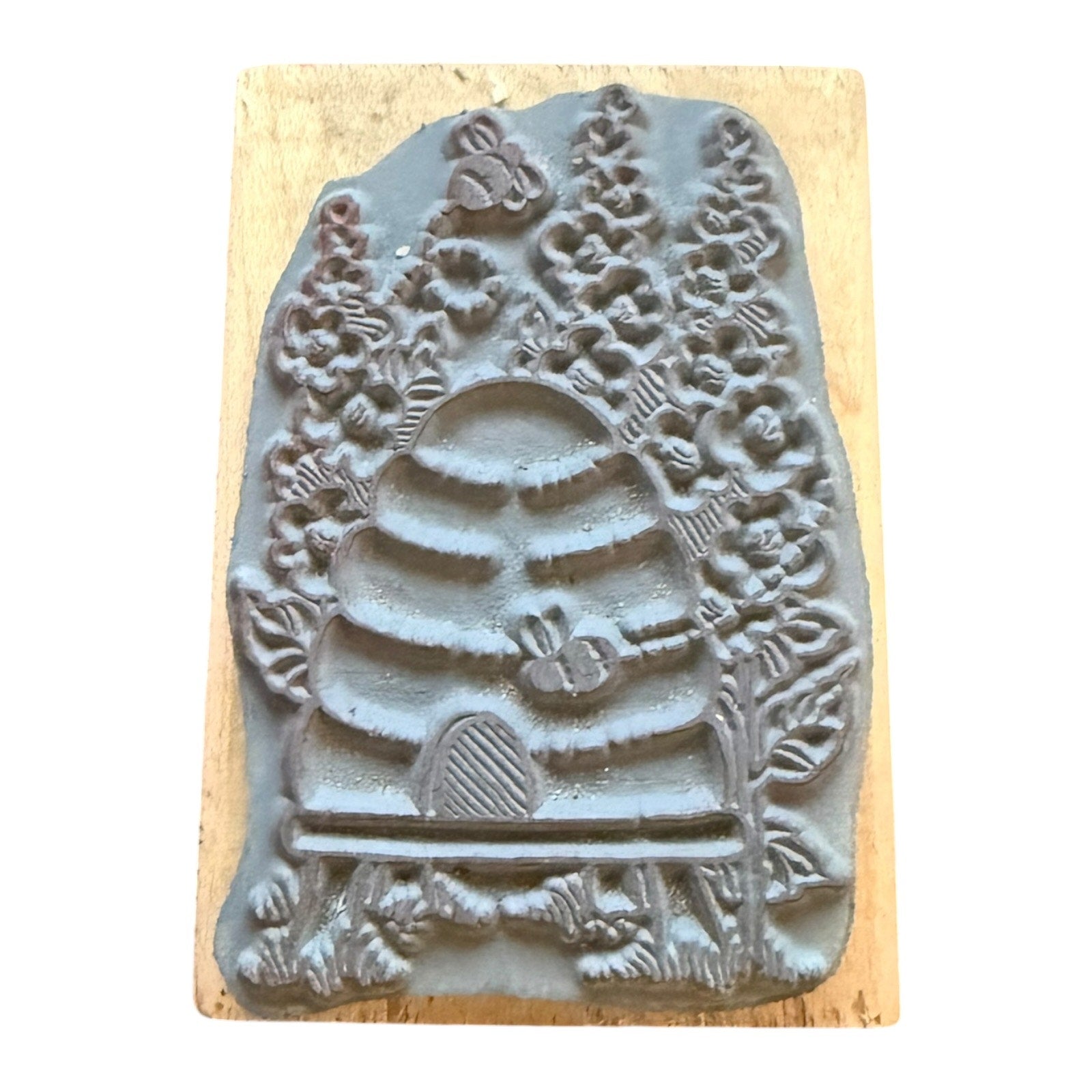 A gray rubber stamp featuring a detailed design of a beehive, bees, flowers, and leaves, mounted on a rectangular light wood block against a white background.