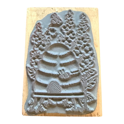 A gray rubber stamp featuring a detailed design of a beehive, bees, flowers, and leaves, mounted on a rectangular light wood block against a white background.