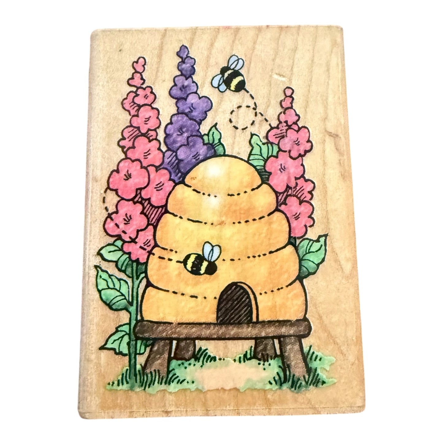 A rectangular wooden stamp featuring a colorful illustration of a beehive, two bees, pink flowers, purple flowers, and green leaves on a light wood background.