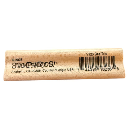 A close-up of a wooden stamp showing the side with printed text including 'Stampenous!', a barcode, and 'V123 Bee Trio' on a plain white background.