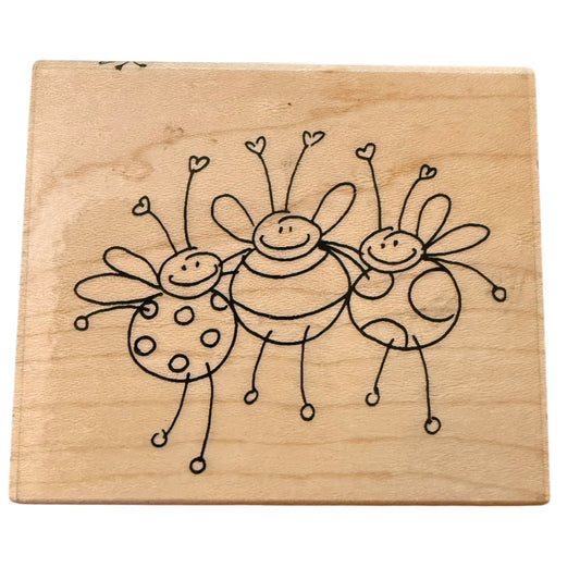A square wood stamp featuring a simple  drawing of three smiling bees on a light natural wood background, shown against a plain white backdrop.