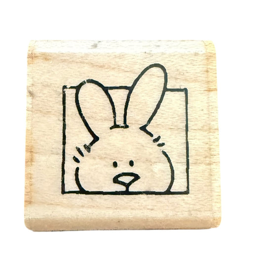 A square wooden rubber stamp block featuring a simple black line drawing of a bunny face peeking from inside a square outline, shown on a plain white background.