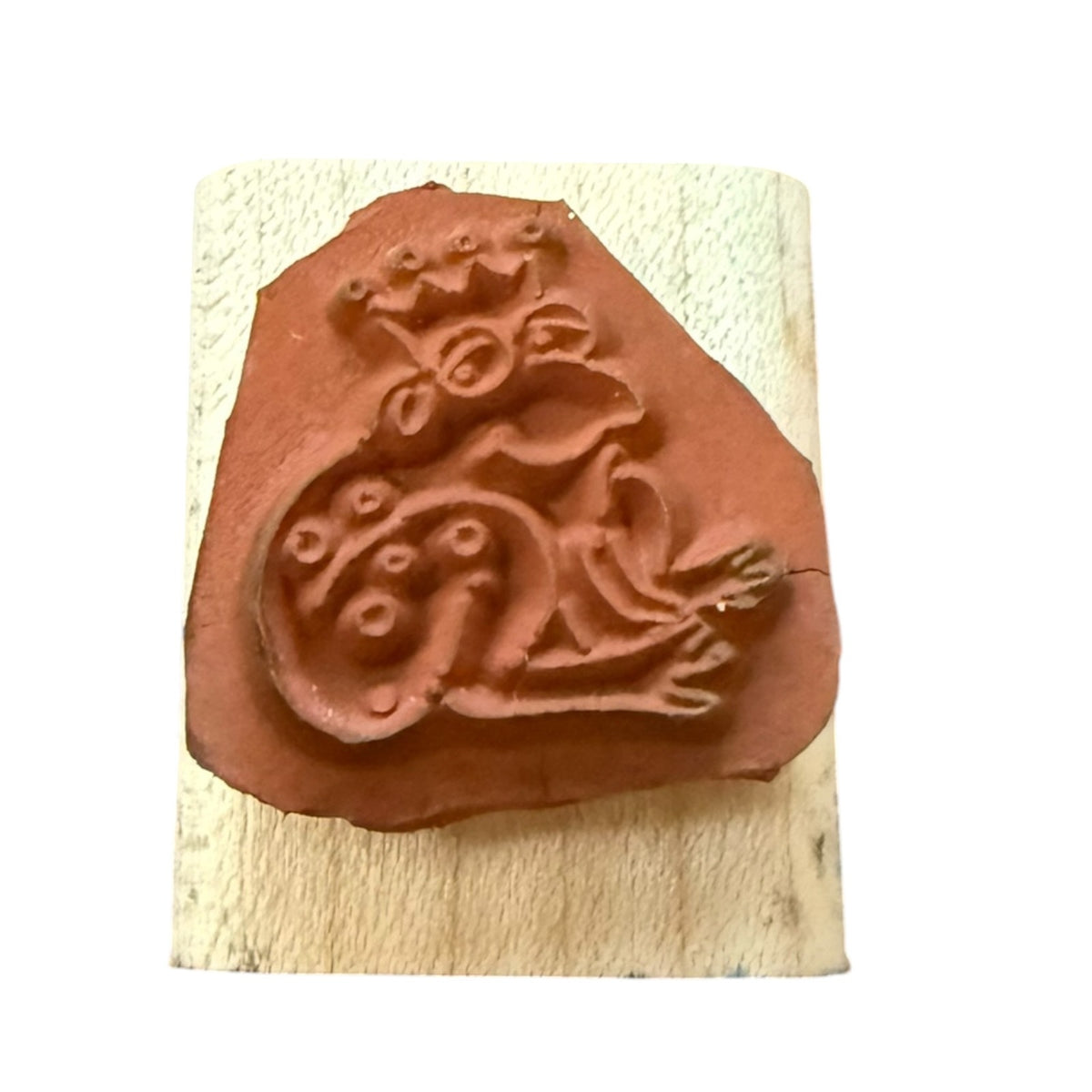 A terracotta-colored stamp featuring a stylized frog design, mounted on a rectangular light wood block against a plain white background.