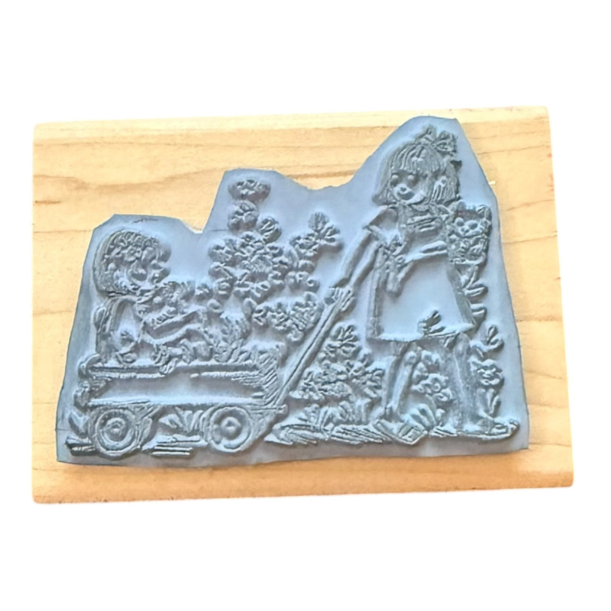 A gray rubber stamp with an embossed illustration of a girl pushing a wagon, mounted on a light wood block against a white background.