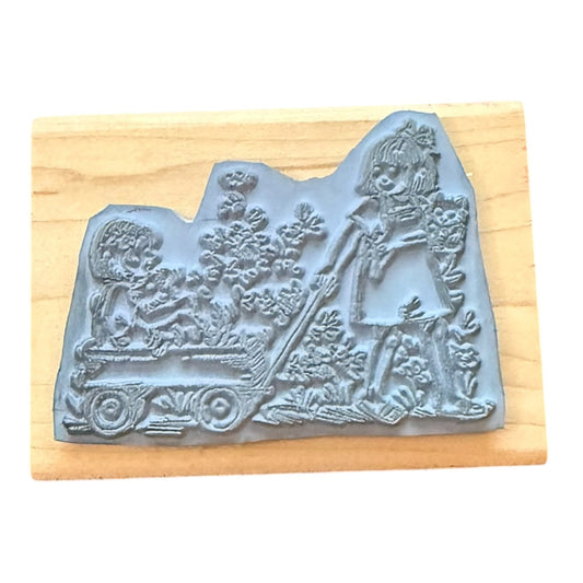 A gray rubber stamp with an embossed illustration of a girl pushing a wagon, mounted on a light wood block against a white background.