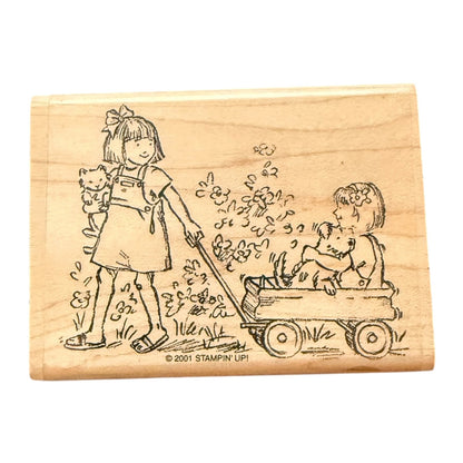 A wooden rubber stamp block featuring an illustration of a girl pulling a wagon with a child & dog sitting inside, plus a small cat, on a plain white background.