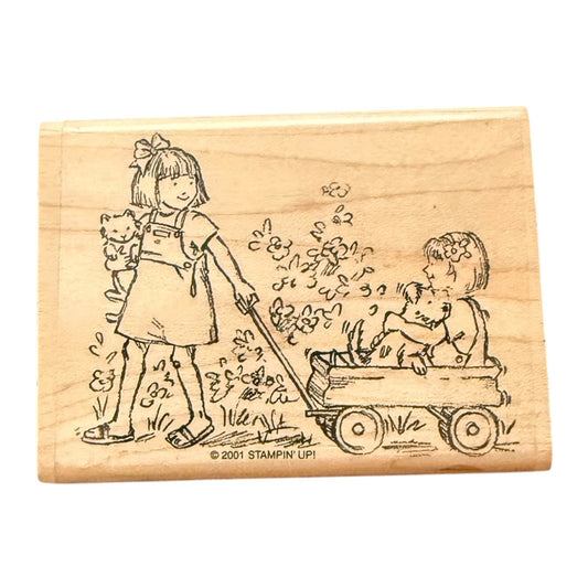 A wooden rubber stamp block featuring an illustration of a girl pulling a wagon with a child & dog sitting inside, plus a small cat, on a plain white background.