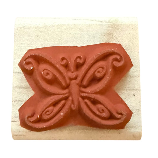 A butterfly-shaped rubber stamp with ornate scrollwork on a light beige wooden block, isolated on a white background.