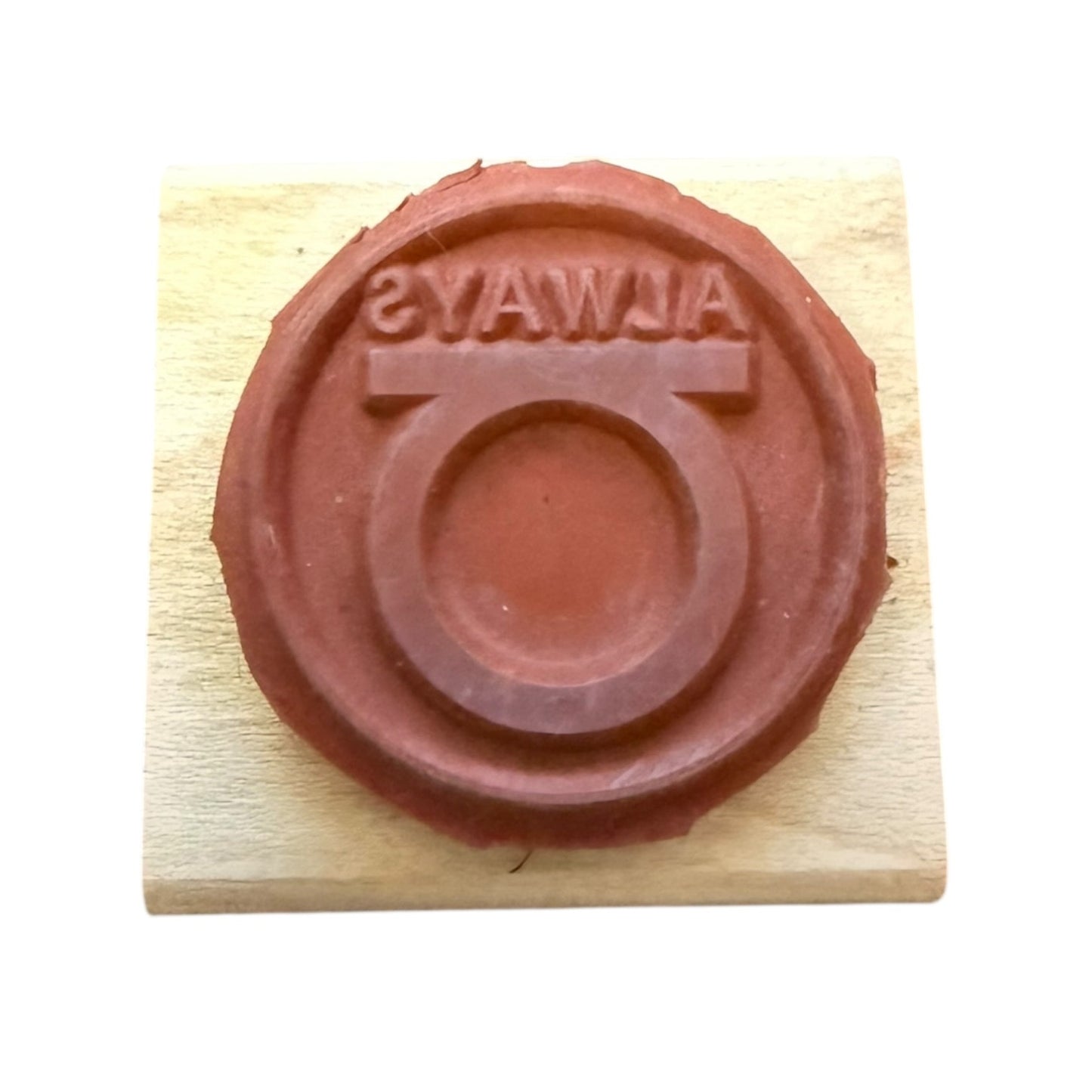 A circular red rubber stamp on a light wood block, featuring the word 'ALWAYS' in reverse around a circular emblem.