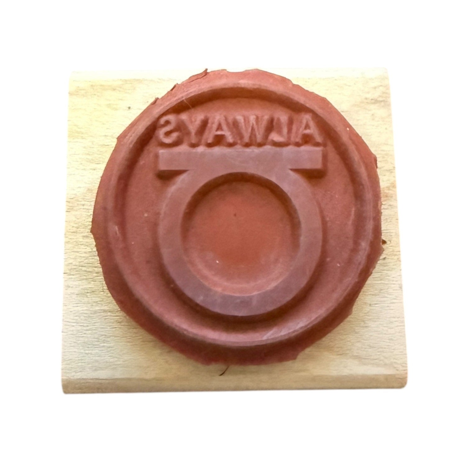 A circular red rubber stamp on a light wood block, featuring the word 'ALWAYS' in reverse around a circular emblem.