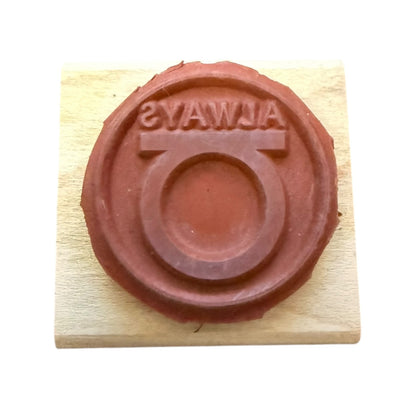 A circular red rubber stamp on a light wood block, featuring the word 'ALWAYS' in reverse around a circular emblem.