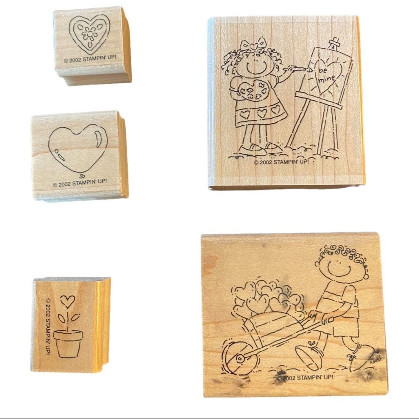 Five wooden rubber stamps shown on a white background, including two larger stamps with engraved line drawings of a girl painting a heart and a boy pushing a wheelbarrow of hearts, and three smaller stamps with simple heart and plant designs. Visible text includes '© 2002 STAMPIN' UP!'.