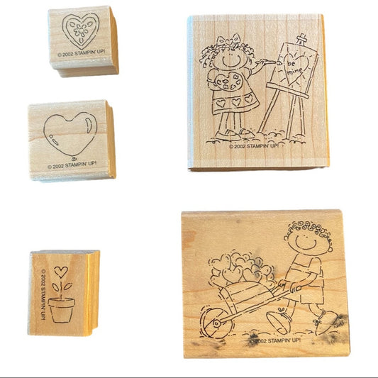 Five wooden rubber stamps shown on a white background, including two larger stamps with engraved line drawings of a girl painting a heart and a boy pushing a wheelbarrow of hearts, and three smaller stamps with simple heart and plant designs. Visible text includes '© 2002 STAMPIN' UP!'.