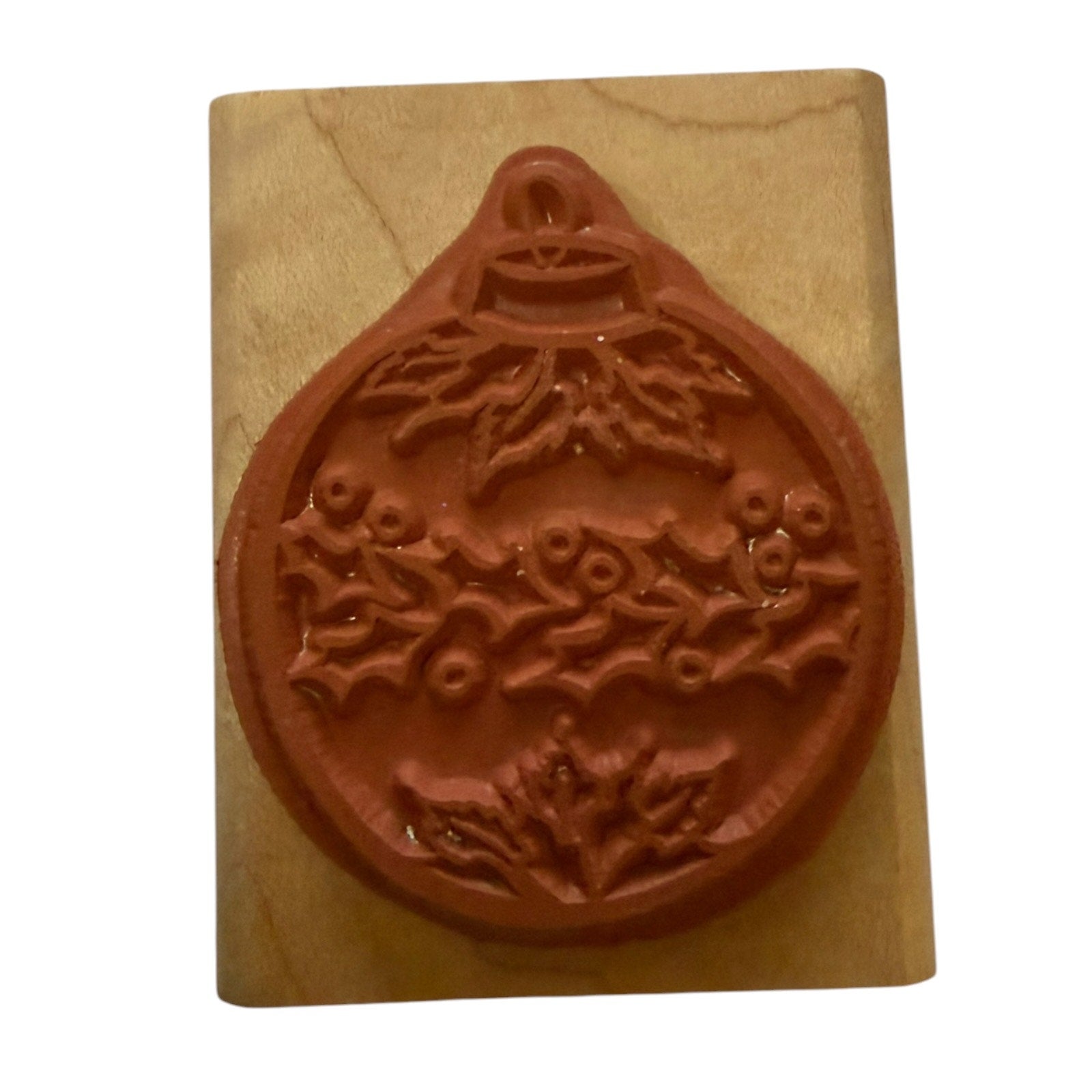 A Christmas ornament-shaped rubber stamp with a festive holly and berry pattern, mounted on a light-colored wooden block against a white background.