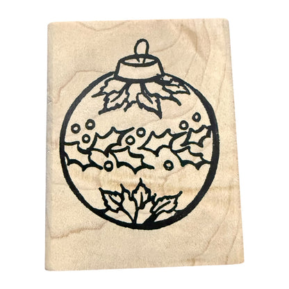 A wooden rubber stamp featuring a black line-art design of a Christmas ornament decorated with holly leaves and berries, shown on a plain white background.