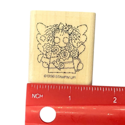 A wooden rubber stamp featuring a black line art illustration of a rag doll girl with flowers in her hair and the text '©1996 STAMPIN UP!', shown with a red ruler for scale on a white background.