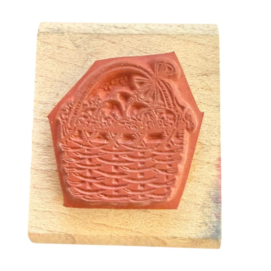 A rubber stamp featuring a basket with flowers and a bow, mounted on a light-colored wooden block against a white background.