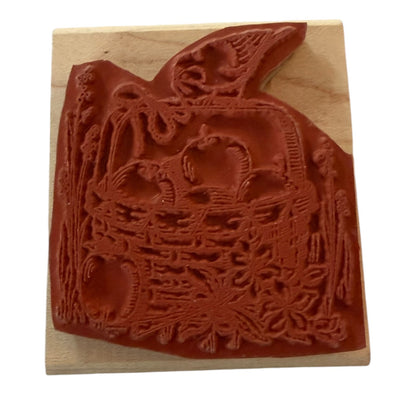 A wooden rubber stamp featuring an embossed autumn harvest design with apple, a basket, and a bird, shown on a light wood block against a white background.