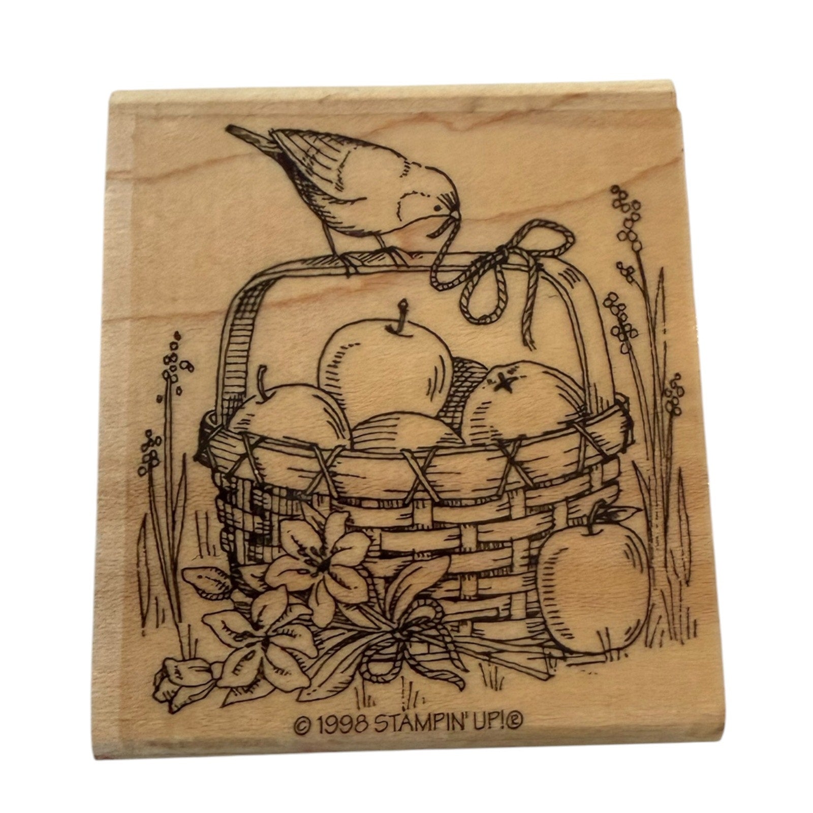 A square wooden stamp block featuring a design of a bird perched on a basket filled with apples, with floral details and the text '© 1998 STAMPIN' UP!' at the bottom.