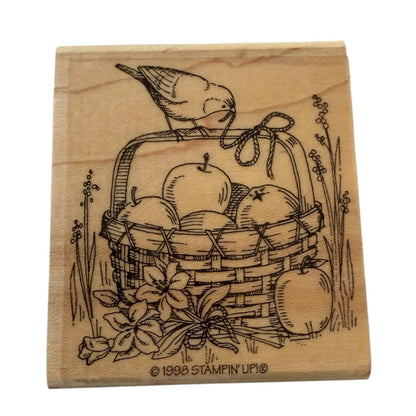 A square wooden stamp block featuring a design of a bird perched on a basket filled with apples, with floral details and the text '© 1998 STAMPIN' UP!' at the bottom.