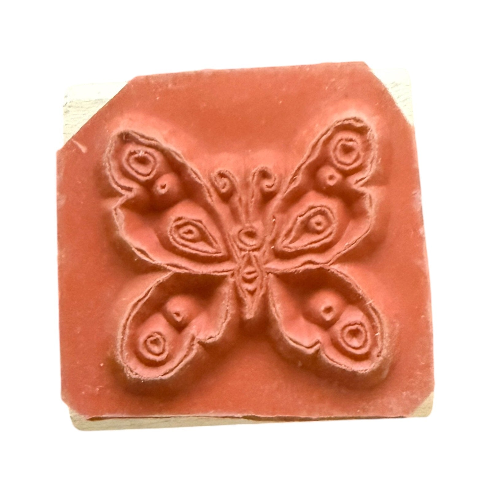 A terracotta-colored rubber stamp with a raised butterfly design, mounted on a light-colored wooden block against a plain white background.