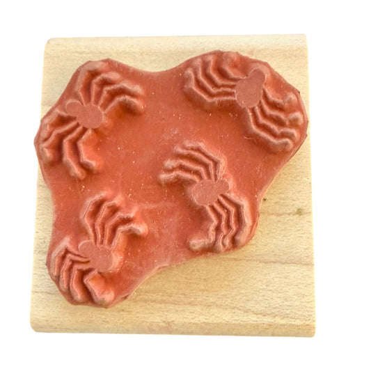 A terracotta-colored rubber stamp featuring a spider design, mounted on a square light wood block against a plain white background.