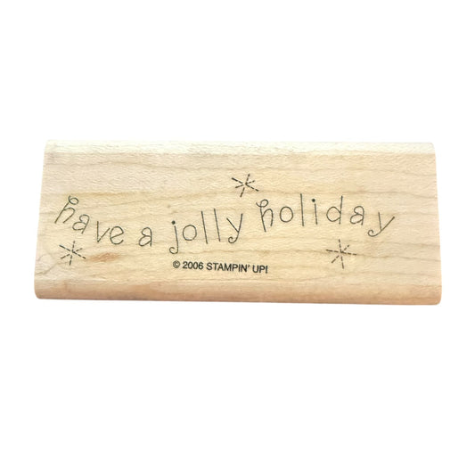 A rectangular wooden rubber stamp with the phrase 'Have a jolly holiday' and small starburst motifs engraved on its light-colored wood surface. The copyright text '© 2006 STAMPIN' UPI' is visible, indicating the brand Stampin' UPI.
