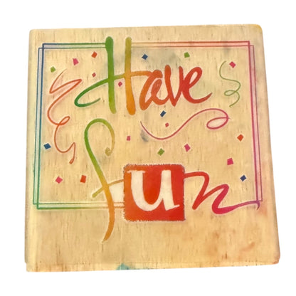 A square wooden block stamp with the words 'Have fun' decorated with small confetti and swirls, on a plain white background.