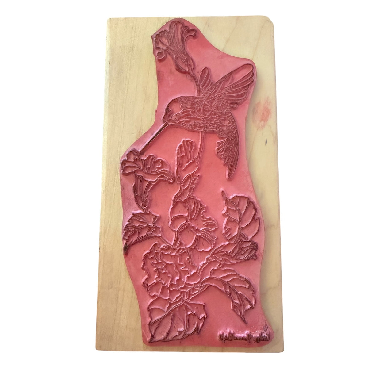 A pink rubber stamp with an embossed design of a hummingbird and flowers, mounted on a light-colored wooden block against a white background.