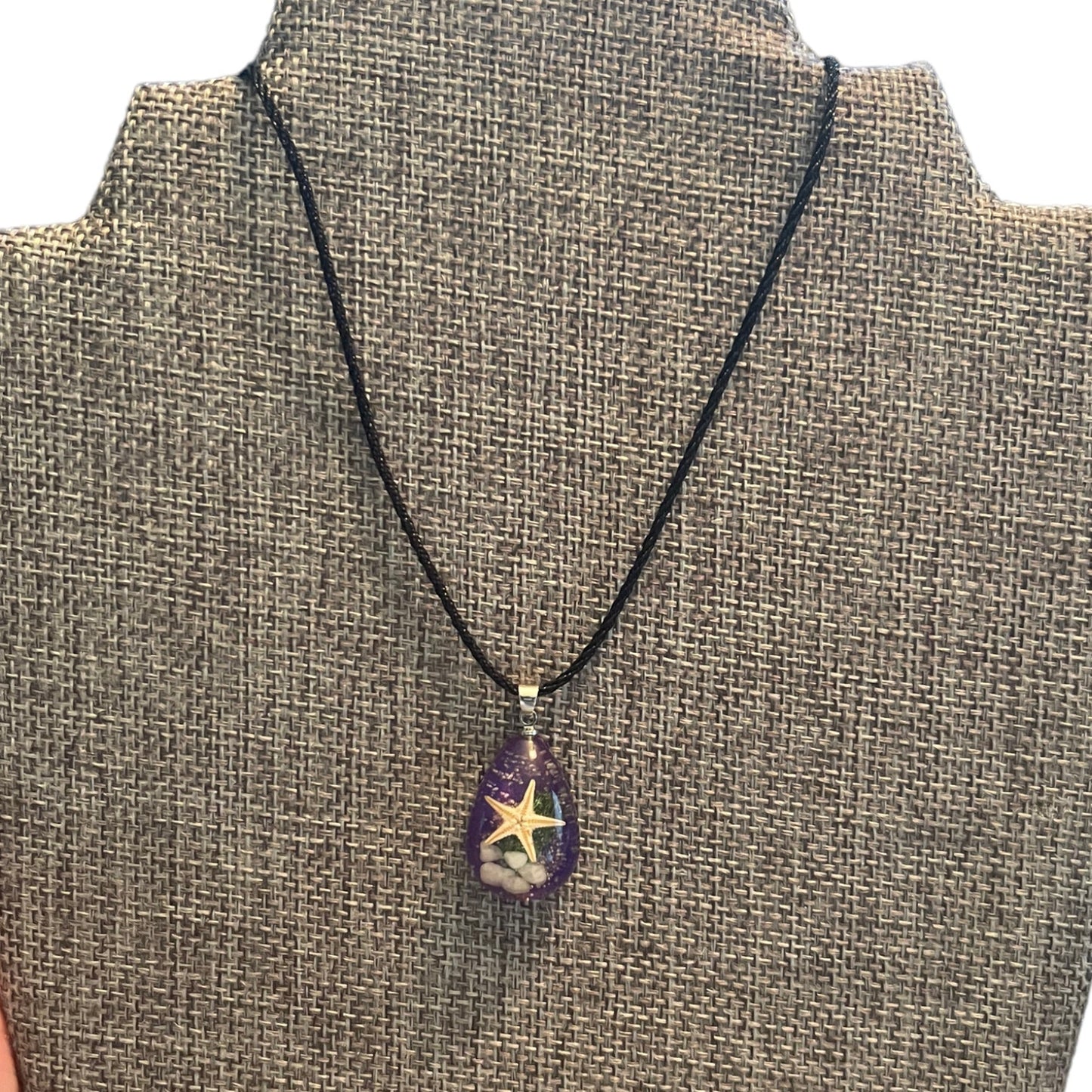 A black cord necklace with a teardrop-shaped purple pendant featuring a starfish design, displayed on a burlap-covered jewelry bust.