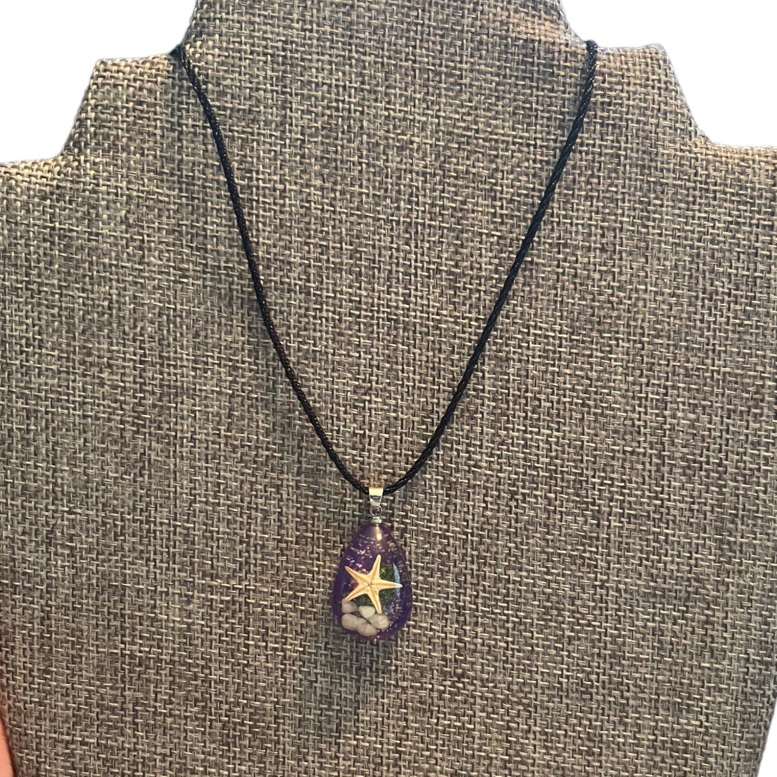 A black cord necklace with a teardrop-shaped purple pendant featuring a starfish design, displayed on a burlap-covered jewelry bust.