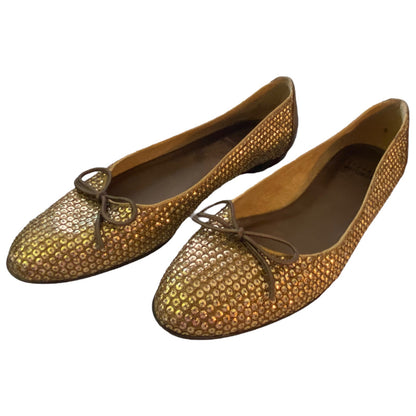 A pair of women's flats covered in small metallic gold sequins with a textured pattern, featuring thin brown leather bows on the toes, shown against a plain white background.